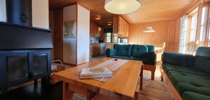 A cozy cabin interior with a wood-burning stove, a green sofa, a wooden dining table, and a kitchen area. - Sauna | Wi-Fi | Terrace | Wood stove | Cable TV (Lofsdalen)