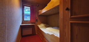 Ein schmales Zimmer mit einem Etagenbett, einem kleinen Schreibtisch mit Stuhl und einem Fenster mit Blick auf ein Gebäude.