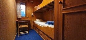 Ein Mehrbettzimmer mit einem Holzbett, einem kleinen Holztisch mit Stuhl, einem Fenster mit Blick auf Bäume und einem blauen Teppich.