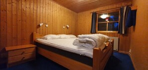 A wooden-paneled room with a bed, bedside table, lamp, and window with curtains.