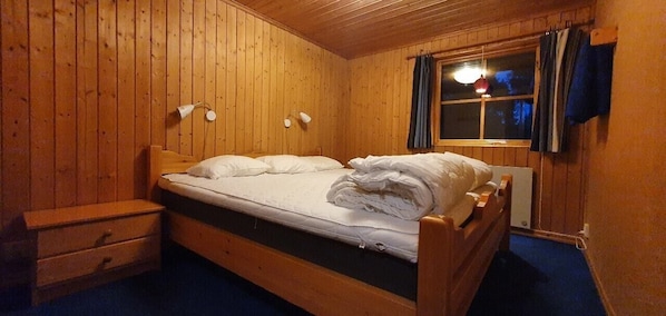 Ein Zimmer mit Holzwänden, einem Bett, einem Nachttisch, einer Lampe und einem Fenster mit Vorhängen.