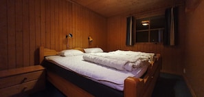 Ein Zimmer mit Holzwänden, einem Einzelbett, zwei Nachttischlampen, einem Fenster mit Vorhängen und einem kleinen Tisch.