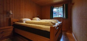 Ein Holzhütten-Zimmer mit einem Einzelbett, einem Nachttisch, einer Lampe und einem Fenster mit Vorhängen.