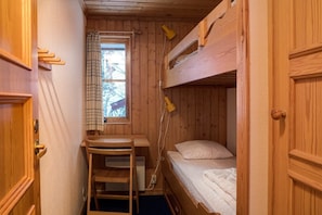 A small, wooden cabin-style room with a bunk bed, a desk, and a window with a curtain.