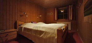 A wooden-paneled room with a bed, a nightstand, a window with curtains, and a wall-mounted lamp.
