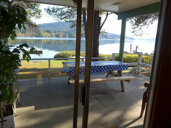 Un porche techado con una mesa y un banco, con vistas a un lago y colinas boscosas.
