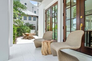 Eine überdachte Terrasse mit Korbmöbeln und einem Holztisch, mit Blick auf einen Garten und ein Gebäude mit großen Fenstern.