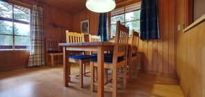 Ein Esszimmer mit Holzboden, einem Tisch und Stühlen, großen Fenstern mit Vorhängen und einer Deckenleuchte.