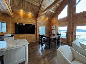 Interior de uma cabana de madeira com área de jantar, TV fixada na parede e grandes janelas com vista para o exterior.