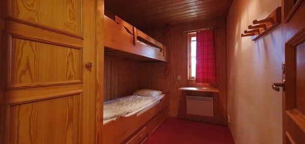 Ein kleines Zimmer mit einem Stockbett, einem Schreibtisch und einem Fenster mit roten Vorhängen.