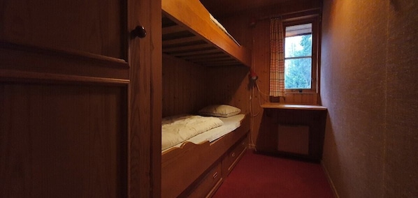 Ein schmales Zimmer mit einem Etagenbett, einem kleinen Tisch und einem Fenster mit Blick auf Bäume.