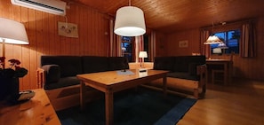 A cozy cabin interior with a wooden dining table, two sofas, a lamp, and a window with curtains.