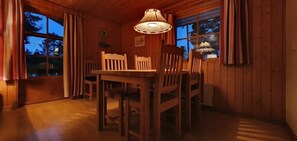A wooden interior with a dining table and chairs, a hanging lamp, and a window with curtains.