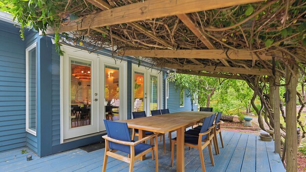 En täckt träveranda med ett bord och stolar, en pergola med klätterväxter och ett hus med blå fasadpanel och en glasdörr.