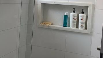 Bruseområde med glasdør, hylde med toiletartikler og brusehoved monteret i loftet.