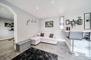 Un salon moderne comprenant un canapé d’angle, un banc, un tapis noir, un coin bar avec des tabourets et des éléments de décoration murale, notamment une citation inspirante et des tableaux encadrés.
