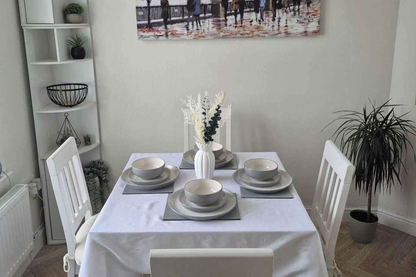 A dining table set with plates and bowls, a vase with white flowers, a shelf with plants, and a painting on the wall.
