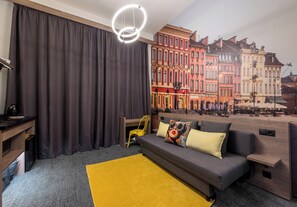 Ein modernes Hotelzimmer mit grauem Sofa, gelbem Teppich und einem Wandbild bunter Gebäude.
