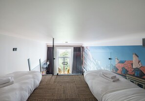 Ein Hotelzimmer mit zwei Betten, Blick auf Gebäude durch ein Fenster und einer Tür, die nach draußen führt.