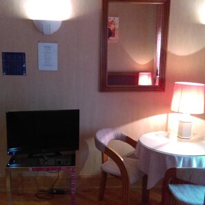 Una habitación con televisión sobre un mueble bajo, una mesa con lámpara, una silla y un espejo en la pared.