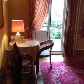 A room with a round table, two chairs, a lamp, a window with curtains, and a view of greenery outside. - Hotel De L Orangerie (Strasbourg)