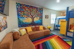 Une pièce décorée d’une grande fresque représentant un arbre, avec un canapé marron agrémenté de coussins, un tapis rayé aux couleurs vives et un lit superposé avec une literie bleue et jaune.