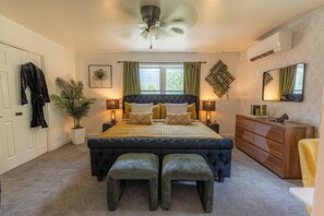 Une chambre à coucher avec une tête de lit capitonnée, une grande fenêtre avec des rideaux, un ventilateur de plafond, une table de chevet avec une lampe, une commode et un miroir.