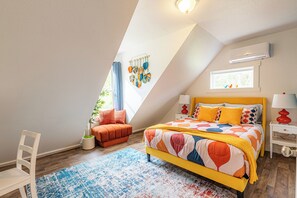 Un dormitorio con techo inclinado, una cama con colcha colorida, una silla naranja pequeña, una mesita de noche blanca con una lámpara, y un aire acondicionado instalado en la pared.