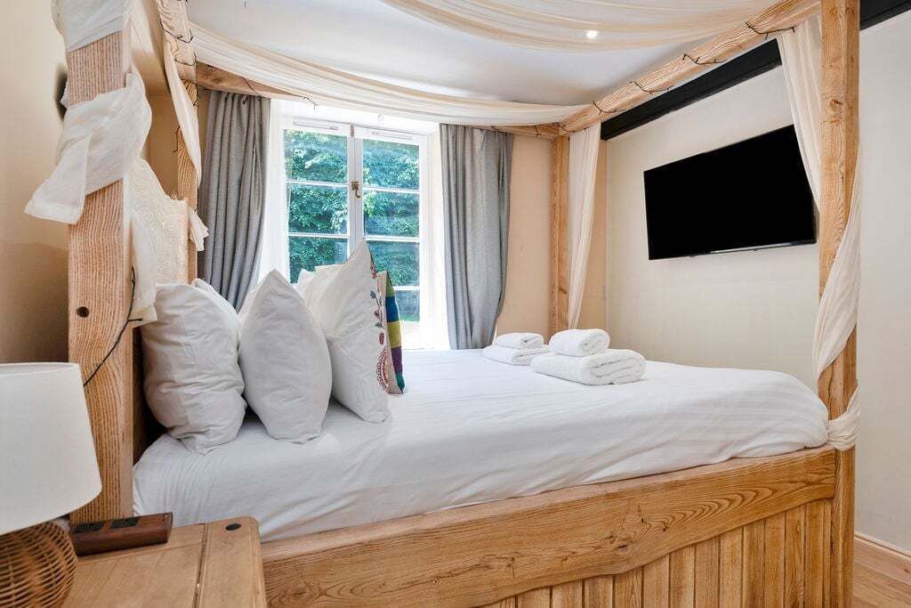 A bedroom with a canopy bed, a flat-screen TV mounted on the wall, and a window with curtains.