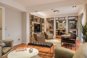 Ein modernes Wohnzimmer mit einer Couch, einem Hocker und einem Couchtisch, ein Essbereich mit Stühlen und einem Tisch sowie ein eingebautes TV-Regal.