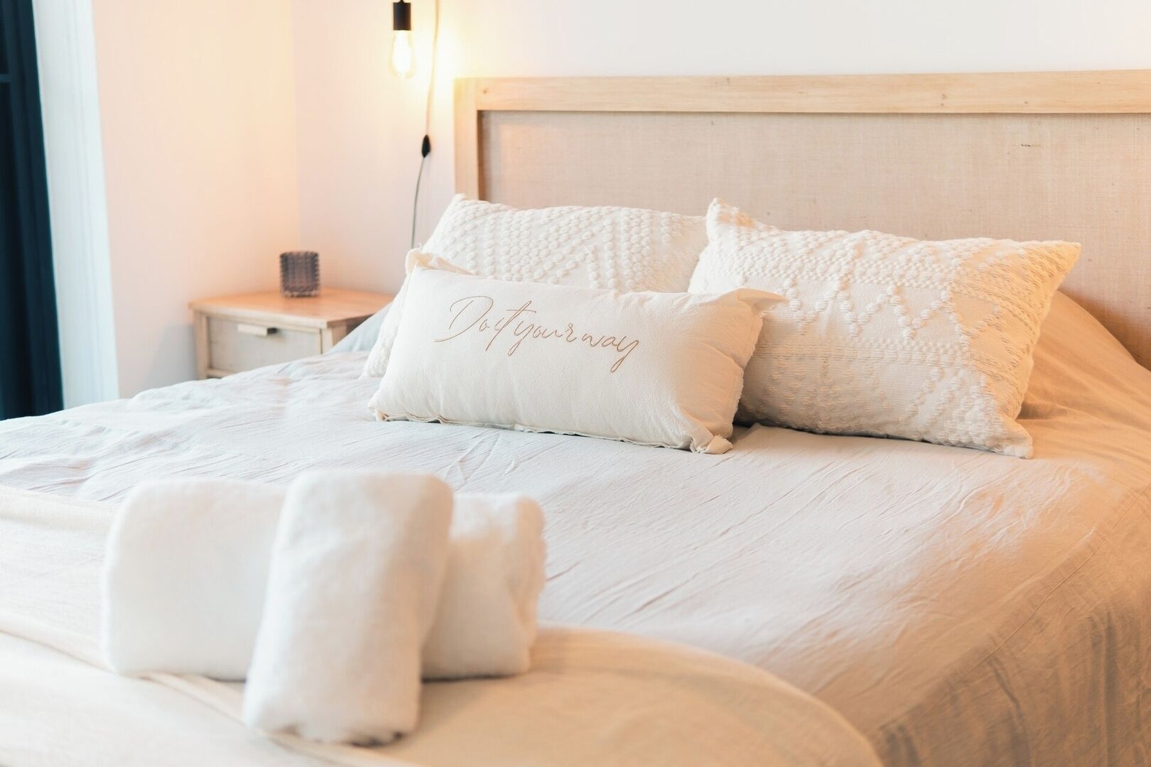 Una cama bien hecha con cabecera, dos almohadas con texto y una mesita de noche con lámpara.