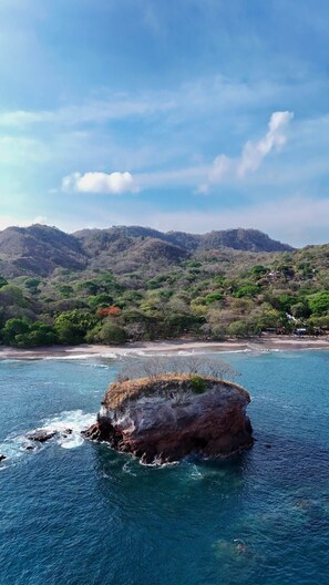 A coastal landscape with a prominent rock formation surrounded by clear blue water, a beach, and a mountainous backdrop. - Villa Të Sula • House of Peace • Coastal Nature Escape (Cabo Velas)
