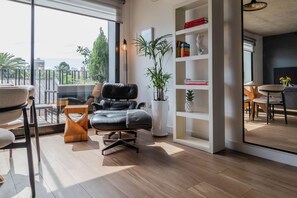 Una sala de estar moderna con un sofá de cuero, una mesa de centro de madera, una estantería y un ventanal con vistas a un paisaje urbano.