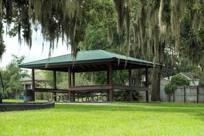 A covered picnic area with benches and a metal roof, surrounded by grass and trees. - The Trail & Tide House, Beaches, Trails, Hikes (Orange Park)