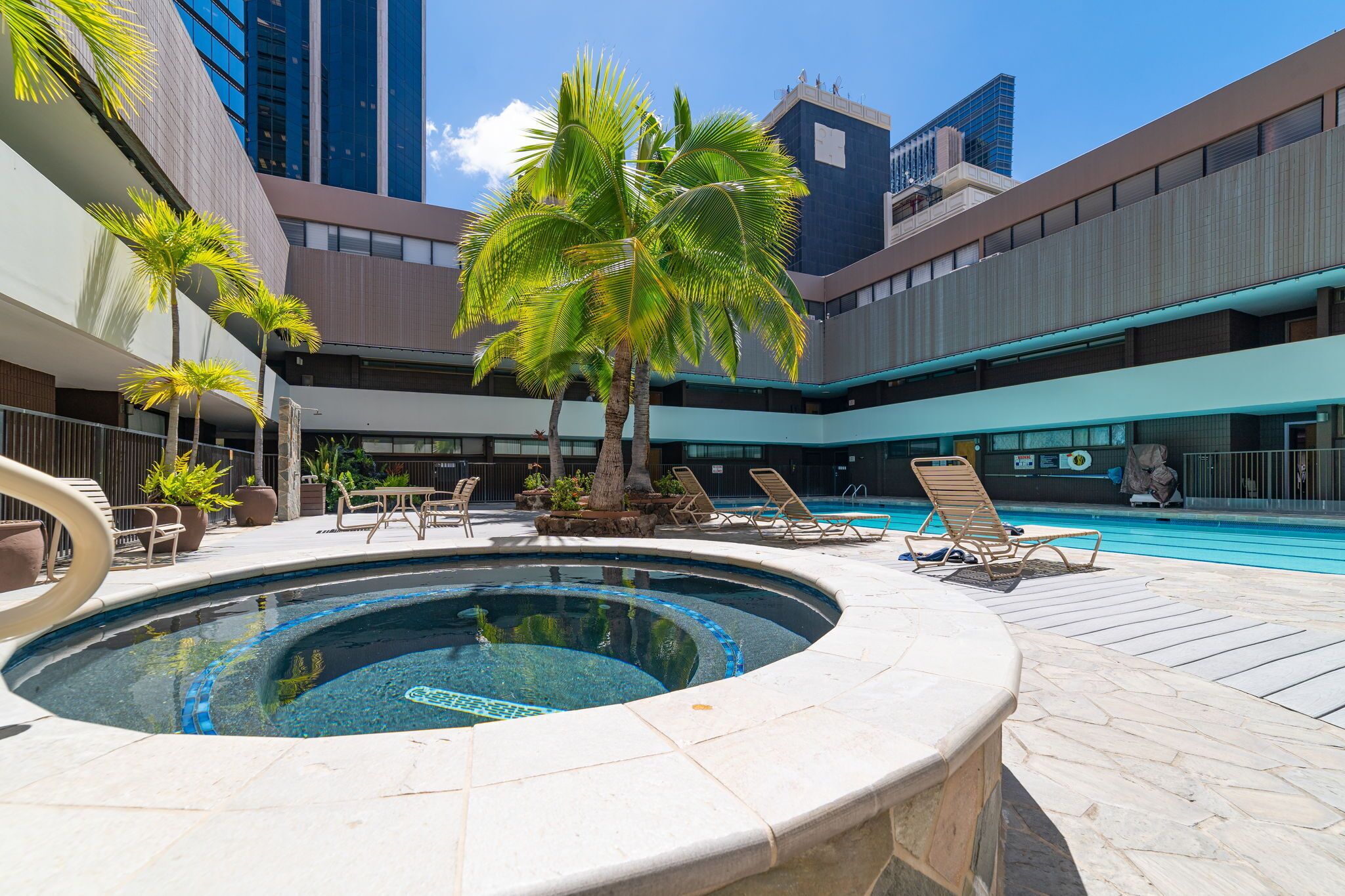 A swimming pool with lounge chairs, palm trees, and modern buildings in the background.. Cozy 1-bedroom apartment with Sofa bed and AC in peaceful Honolulu