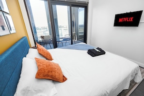 A hotel room with a bed, orange pillows, a TV screen displaying 'NETFLIX,' and a view of the city through large windows.