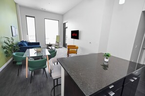 A modern living room with a glass dining table, green and white chairs, a blue sofa, a yellow armchair, and a large window.