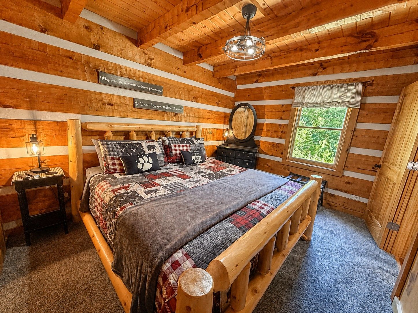 A log cabin bedroom with a wooden bed, a nightstand, a mirror, and a window with a view of greenery.