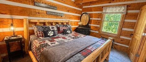 A log cabin bedroom with a wooden bed, a nightstand, a mirror, and a window with a view of greenery.