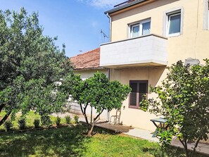 Ein zweistöckiges Gebäude mit Balkon, ein kleiner Garten mit Bäumen und ein klarer Himmel.