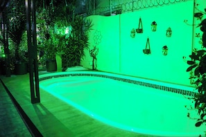 A swimming pool with a green-lit edge, surrounded by potted plants and decorative lanterns on a wall.