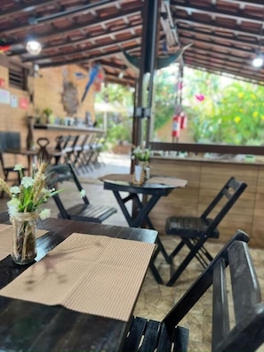 An outdoor seating area with tables and chairs, a wooden ceiling, and a view of greenery.
