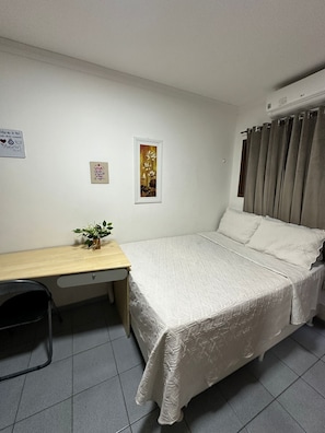 A single-bed room with a wooden desk, a small plant, a framed picture, and a curtain.