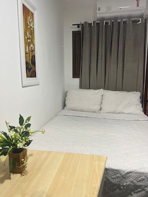 A bedroom with a bed, a wooden table, a potted plant, a painting, and a window with curtains. - Pousada Flor Dália (Natal)
