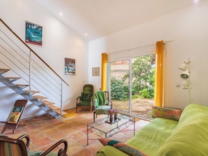 A living room with a green sofa, a wooden coffee table, a green armchair, a staircase with a metal railing, and a large window with yellow curtains. - Vacation home Marmul by Interhome (Hyères)