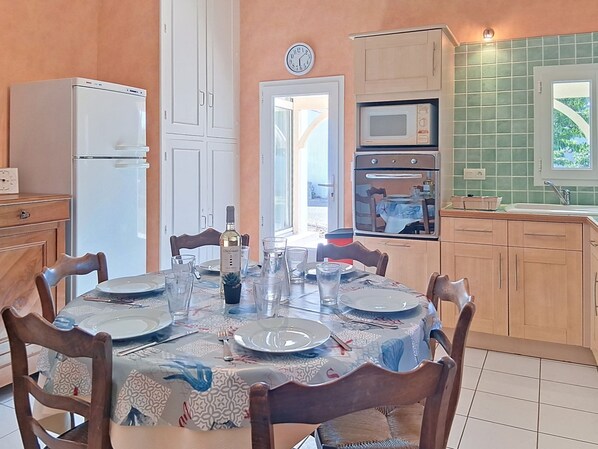 A dining area with a table set for four, a clock on the wall, and a kitchen with a microwave and oven. - Les Tamarix by Interhome (Ile d'Oléron)