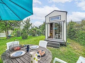 Eine kleine Holzhütte mit einer Schiebetür aus Glas, einem Tisch mit Tellern, Tassen und einer Kanne, sowie einem blauen Sonnenschirm.