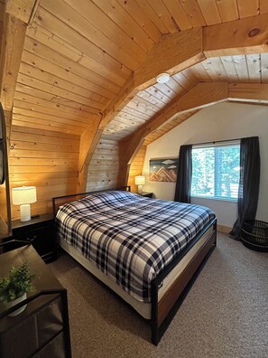 Ein Schlafzimmer mit Holzbalkendecke, einem Bett mit karierter Tagesdecke, einem Nachttisch mit Lampe, einem Fenster mit Vorhängen und einem kleinen Tisch mit Pflanze.