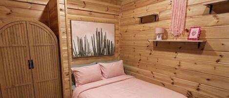 Una habitación con paneles de madera, una cama, un armario, una estantería con objetos decorativos y un atrapasueños colgado en la pared.