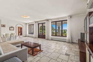 Ein geräumiges Wohnzimmer mit einer Couch, einem Couchtisch und einer TV-Konsole. Große Fenster mit Vorhängen, ein Essbereich mit Stühlen und ein Blick ins Grüne.